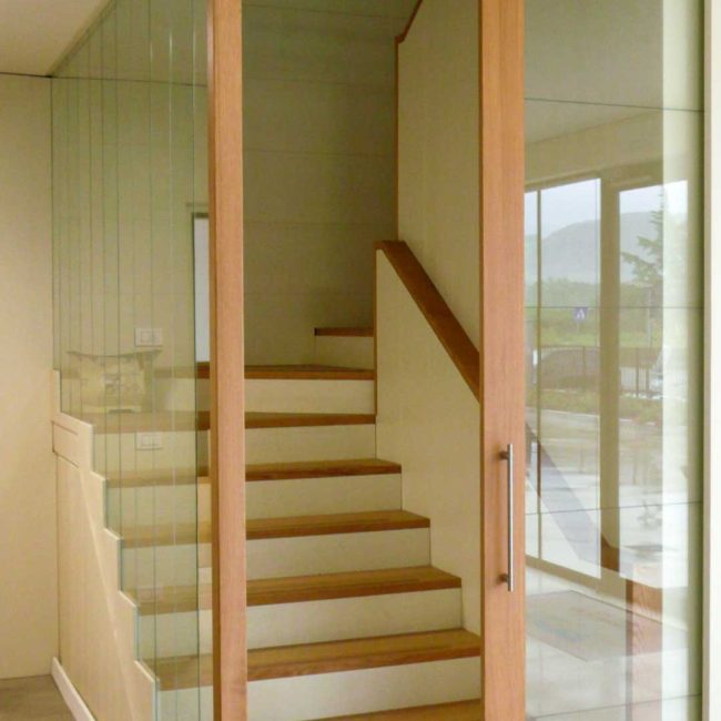 Scala con pedate in rovere e alzate laccate Ral 1013 leggermente spazzolate. Parapetto costituito da fasce di vetro trasparente temperato.