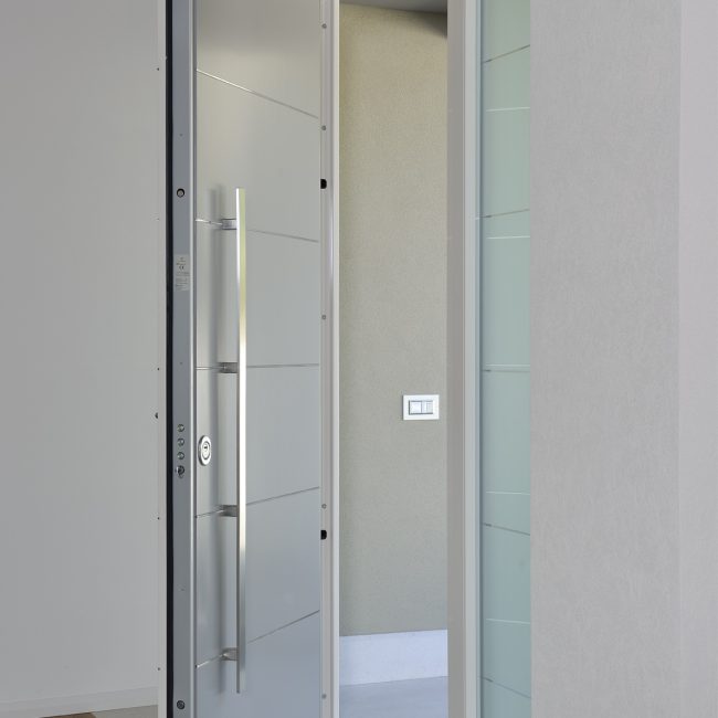 Portoncino blindato laccato con maniglione e inserti in acciaio inox, parti laterali in vetro satinato con righe trasparenti.